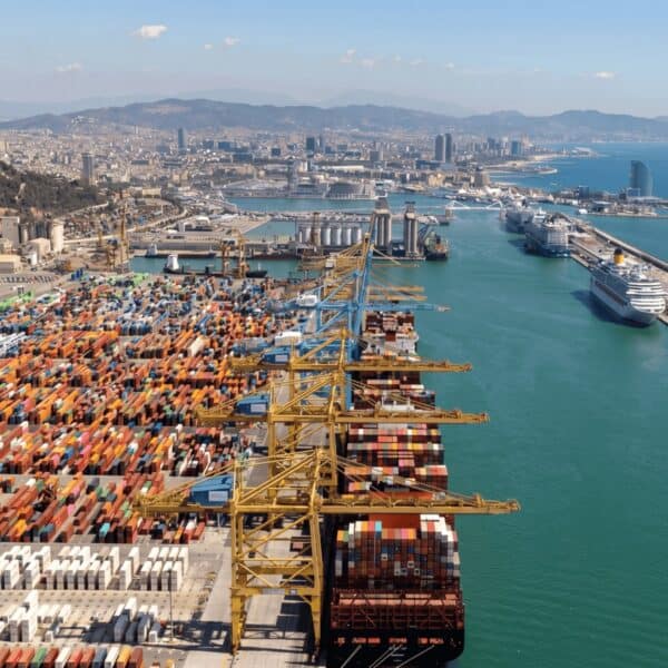 Imagen de un puerto con contenedores apilados, grúas y barcos de carga en un día soleado.